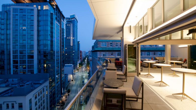 A modern rooftop terrace at dusk with a view of urban high-rise buildings; features outdoor seating and tables, and city lights are visible.