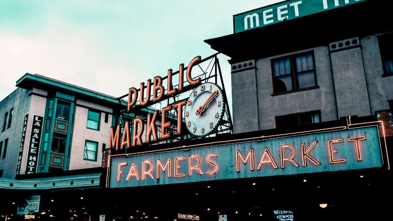 The image shows the iconic neon sign of "Public Market" and "Farmers Market" at Pike Place Market in Seattle, with surrounding buildings visible.
