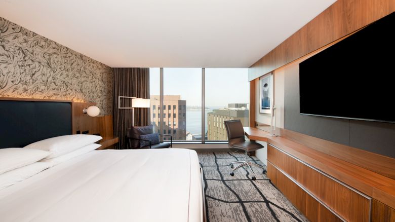 The image shows a modern hotel room with a large bed, a TV, a desk, and a window offering a city view.