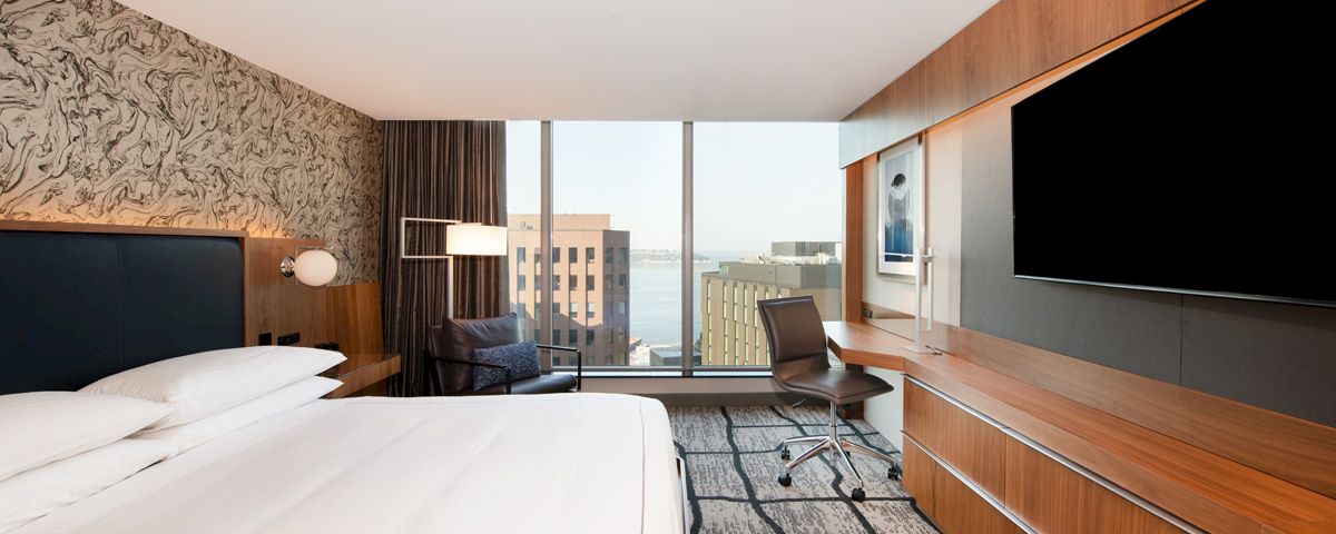 A hotel room with a bed, desk, chair, and TV, and a city view out the window.