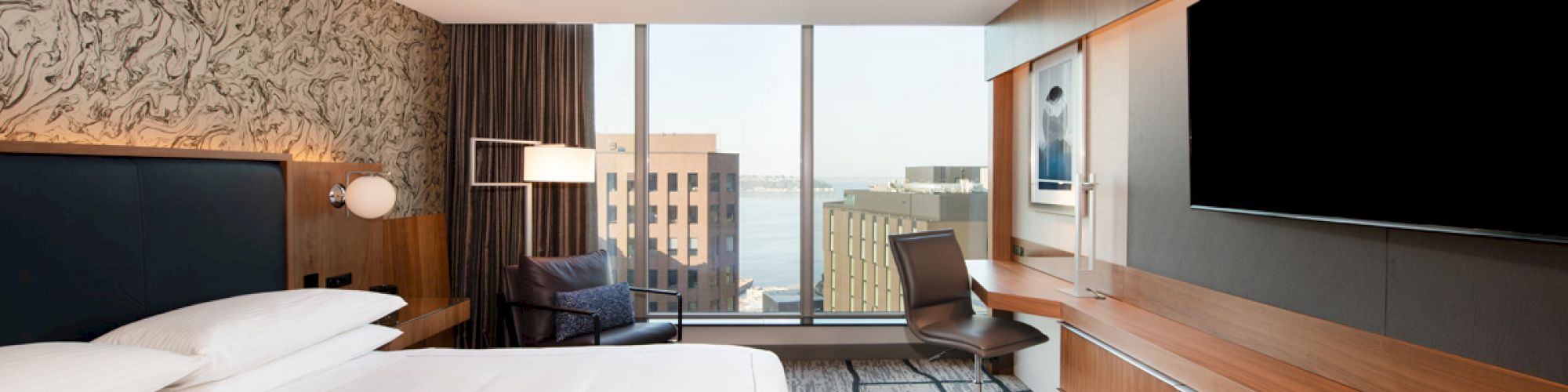 A modern hotel room with a large bed, a desk with a chair, a wall-mounted TV, and a window with a city view is shown.
