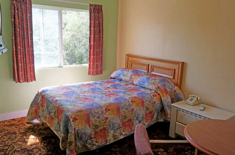 A simple room with a double bed with colorful bedspread, a nightstand, a round table with a chair, curtains, a window, a TV, and a fan.