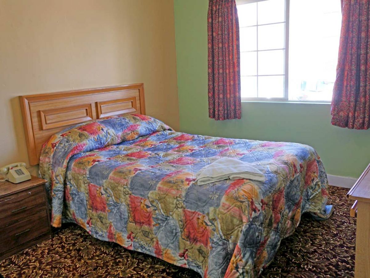 A small bedroom with a colorful patterned bedspread, nightstand with a phone, window with curtains, a TV on a dresser, and carpeted floor.