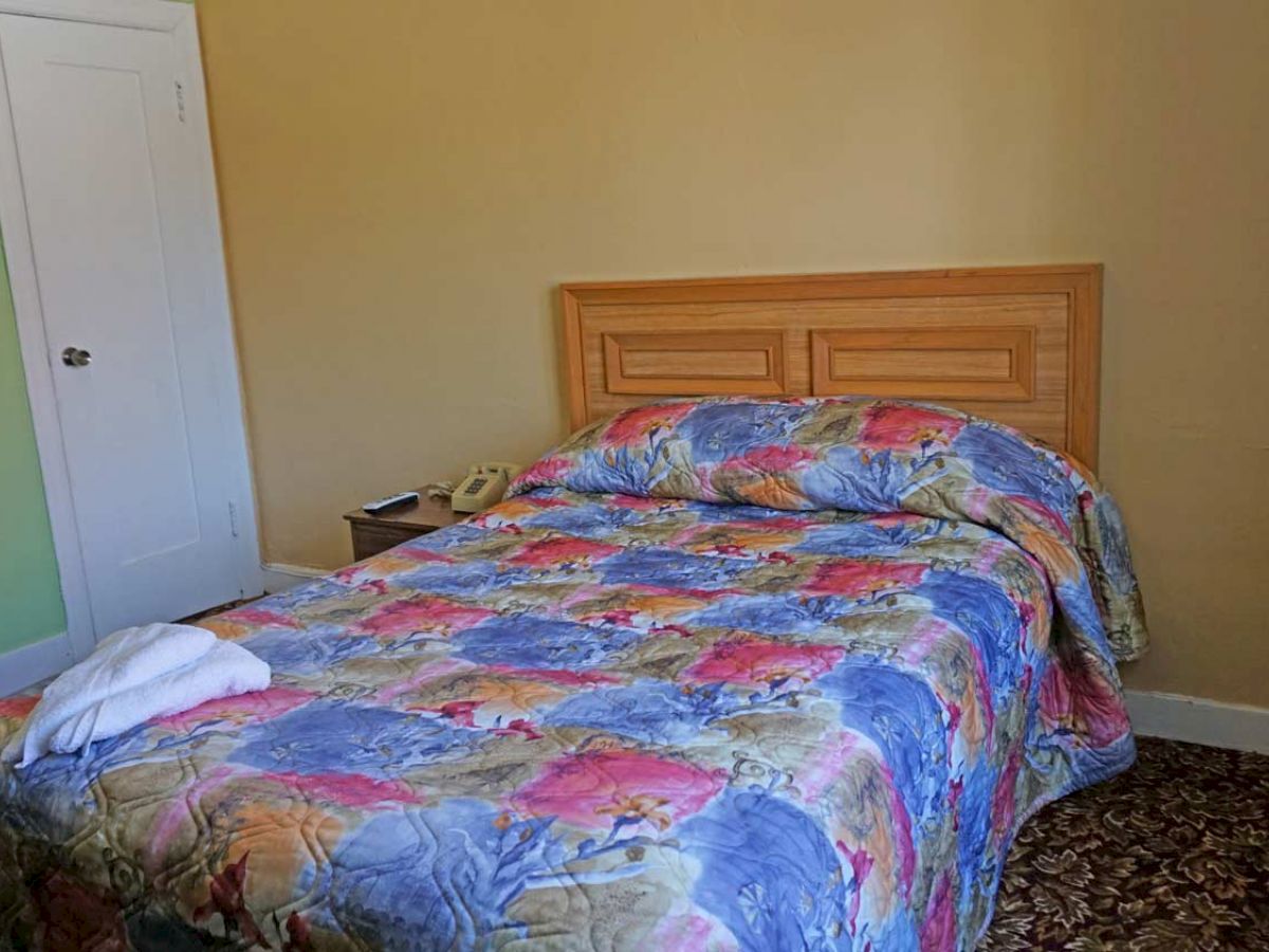 The image shows a neatly made bed with a colorful floral comforter, a nightstand with a telephone, a small white towel on the bed, and a closet door.