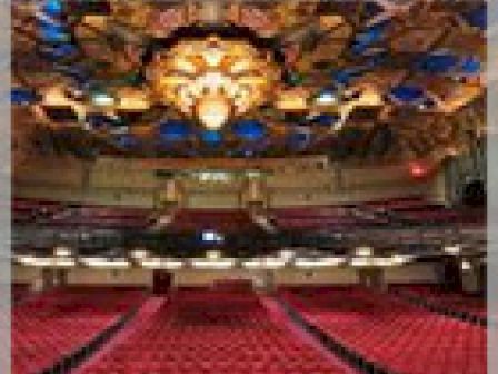 A large, ornate theater with red seats and a decorated ceiling. The stage and proscenium arch are visible, with intricate design elements overhead.