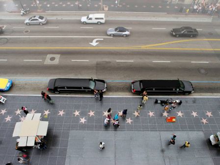 An aerial view of a street with star tiles on the sidewalk, two black limousines, and several people walking.