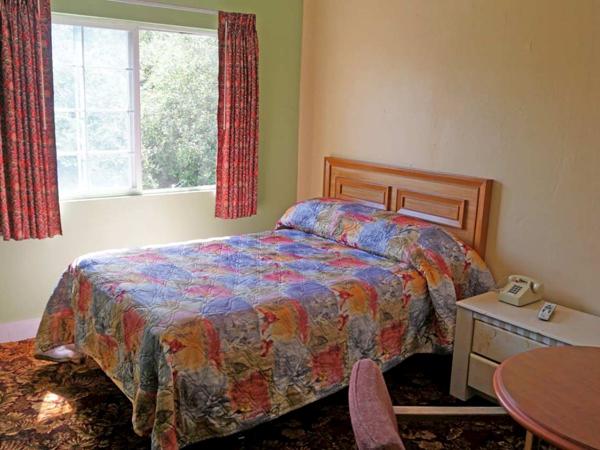 A small bedroom features a bed with a colorful quilt, a window with red curtains, a TV, a fan, a bedside table, a phone, and a round table.